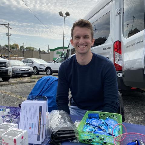 NHC PGH member Brett smiling at a health event outside, behind a table of health education materials