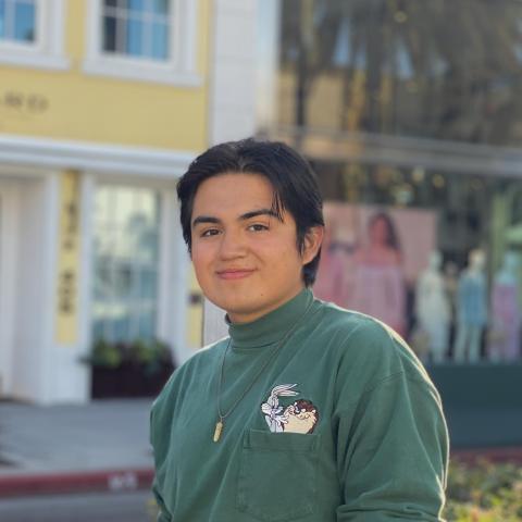 Steven Garcia-Machuca, facing towards the camera while turning to the right, smiling with lips closed. He is wearing an olive green long sleeve t-shirt with the characters Bugs Bunny and the Tasmanian Devil on a pocket on his left chest. Behind him are the entrances to shops, the one on the left with yellow paint and white door and window frames, and the right with pink-red coloring. 