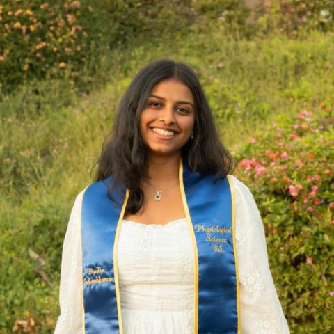 Nivedha stands in the middle of the frame, facing the camera and smiling. She is wearing a white dress and a graduation stole reading "Nivedha Satheeshkumar" and "Psychological Science, B.S.". Behind her is a field of grass and flowers. 