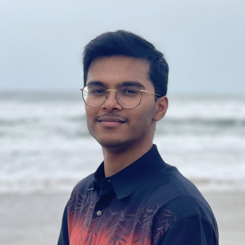 Shreyas Raj is standing in the center of the frame, facing the camera and smiling. Behind him is the ocean with an overcast sky.. 