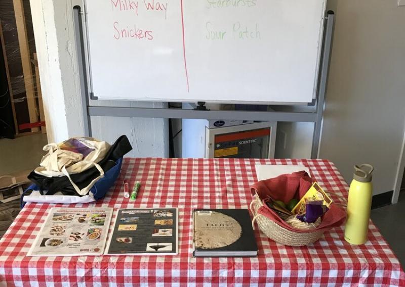 A table at HR 360 Food Pharmacy with a few recipe books, a basket, and a whiteboard behind the table reading "Happy Halloween at HR 360!" and a poll asking which candy has more sugar (Reese's, Milky Way, Snickers, Sour Patch, Starbursts, Red Vines). 