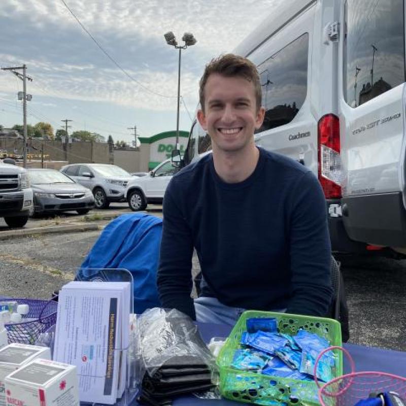 NHC PGH member Brett smiling at a health event outside, behind a table of health education materials