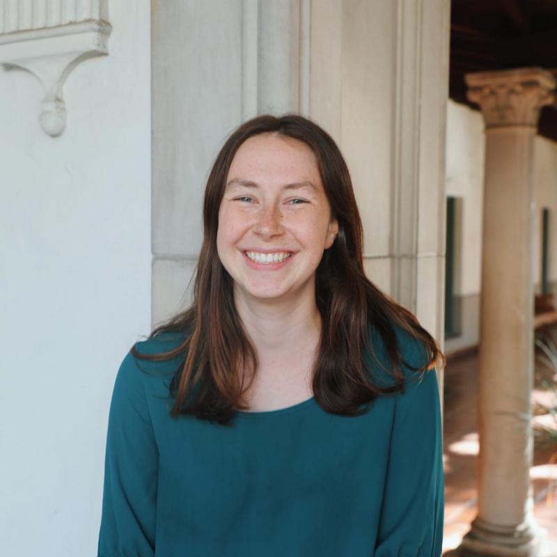 Catherine is standing in the middle frame, facing the camer and smiling. She is wearing a green blouse. Behind her is a white doorframe, and in the back you can see an ionic column.