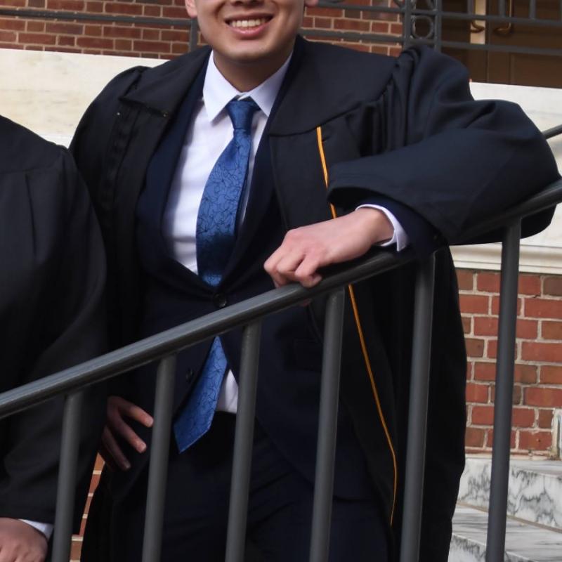 Trevor is standing in the middle of the frame, smiling at the camera. He is wearing square rimmed glasses. He is also wearing black graduation regalia over a navy suit with a white collared shirt and blue tie. 