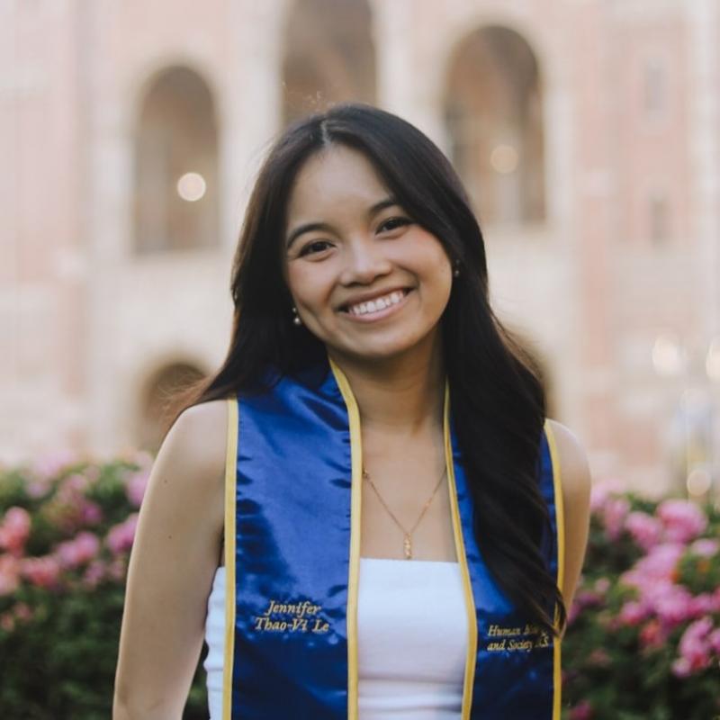 Jennifer is standing in the middle of the frame, facing the camera and smiling. She is wearing a white dress. She has a blue and gold UCLA graduation stole draped over her shoulders. 