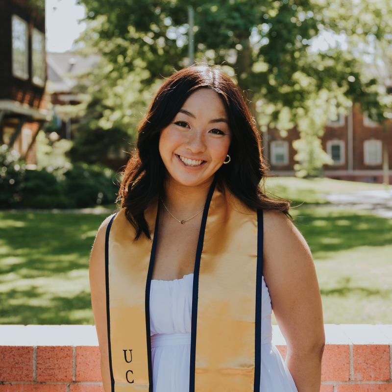 Samantha is standing in the middle of the frame, facing the camera and smiling. She is wearing a white dress. She has a blue and gold UC Davis graduation stole draped over her shoulders. 