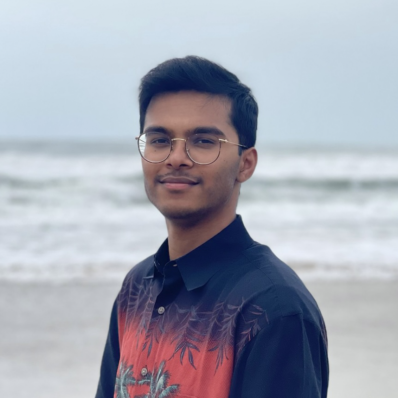 Shreyas is standing in the center of the frame, facing the camera and smiling. Behind him is the ocean with an overcast sky.. 