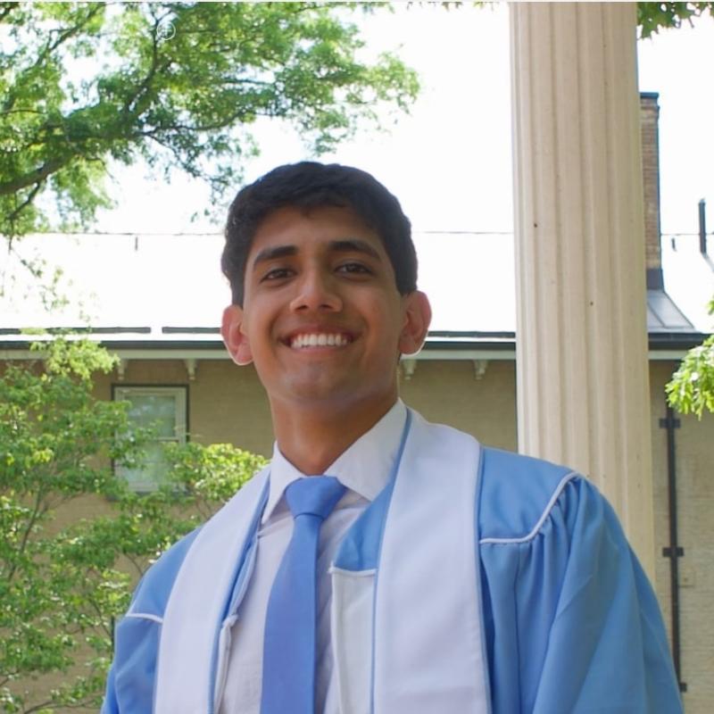 headshot of Dev smiling in blue graduation gown and tie