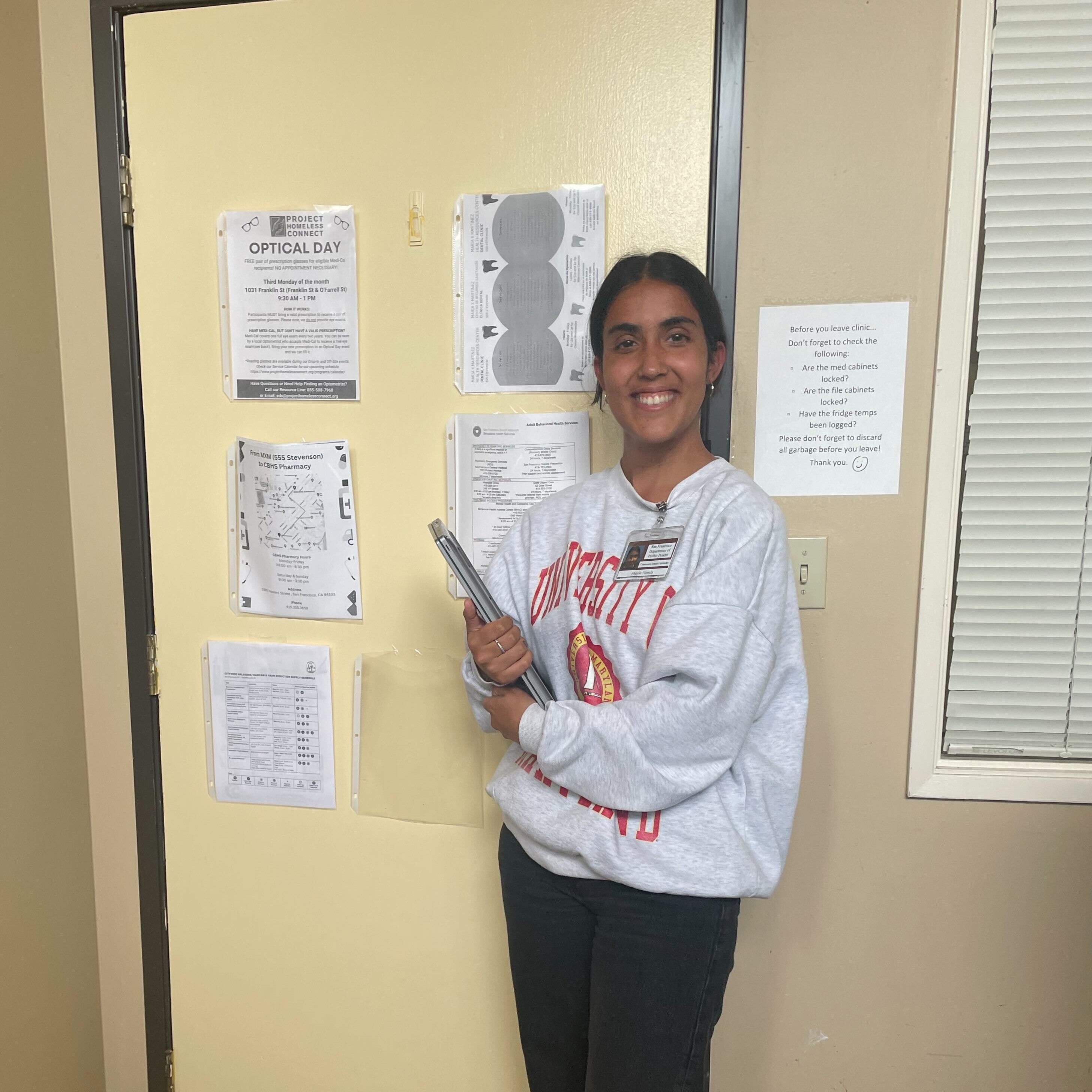 Anjalie is standing off-center, facing the camera and smiling - she is holding her laptop on her way out of the office.