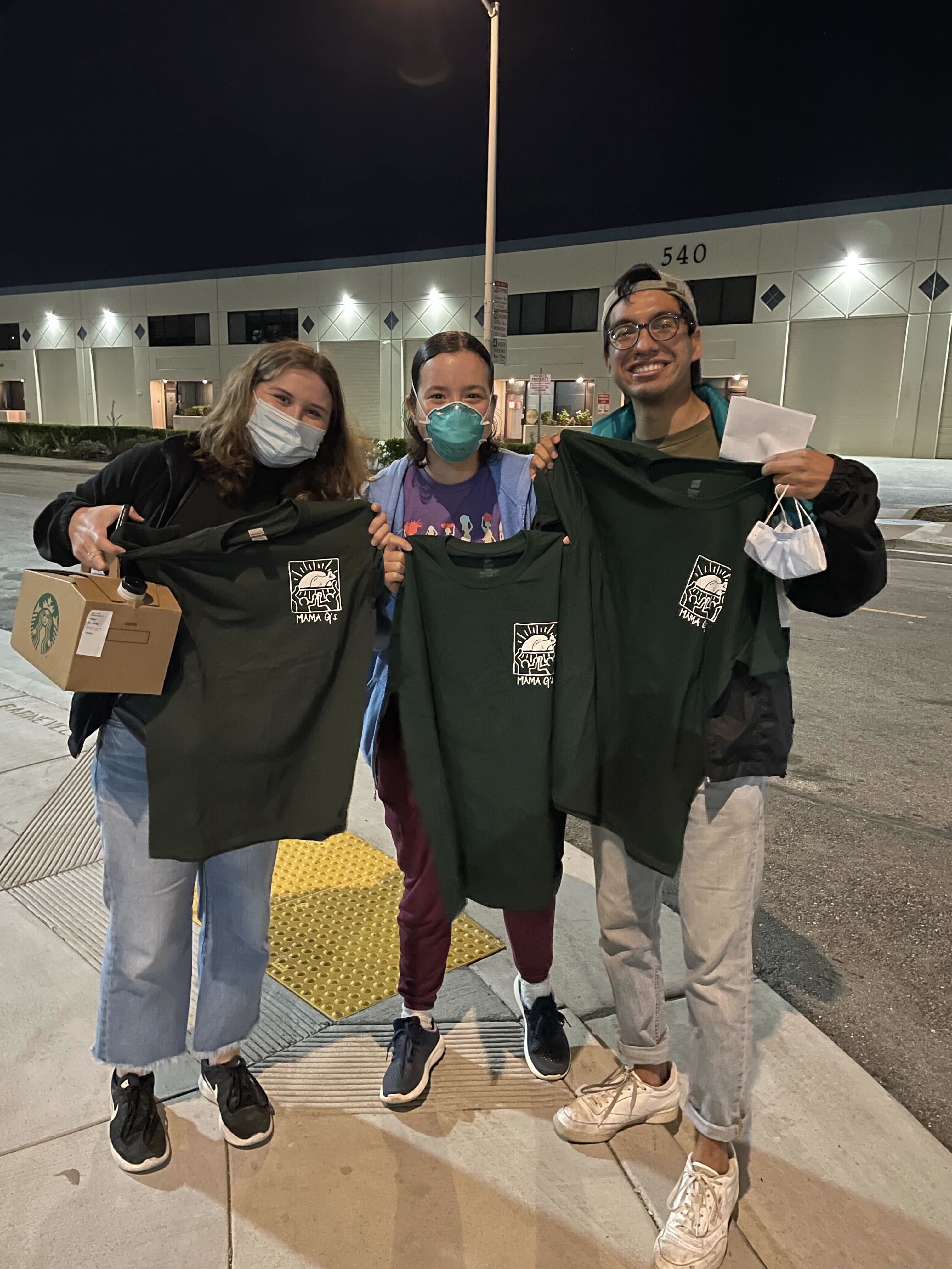 three people stand in the center of the frame facing the camera - each person is holding up a shirt that reads "Mama G's", the name of a non-profit organization in San Francisco.