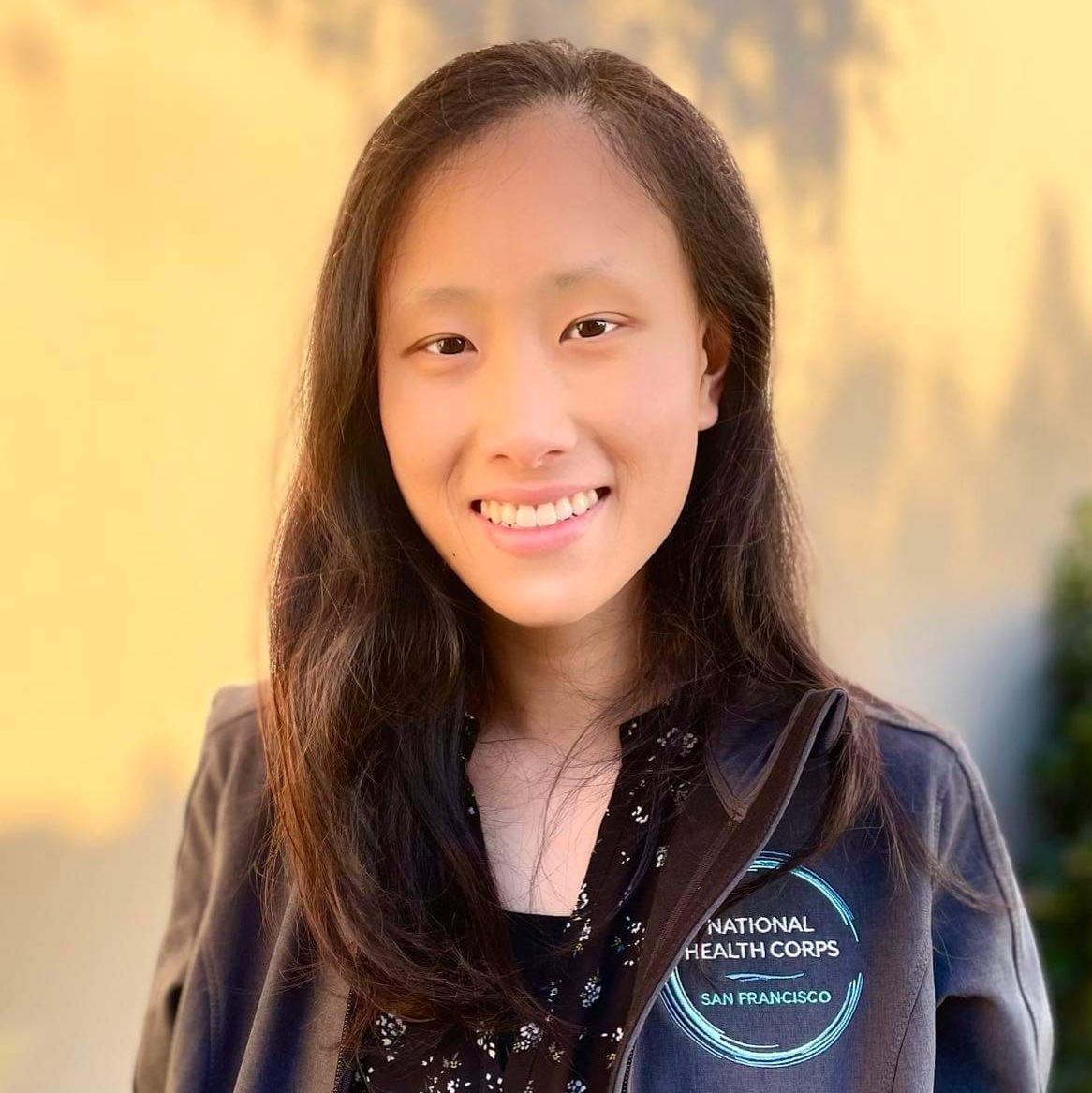 Mindy is seen facing the camera and smiling. She is wearing her charcoal grey National Health Corps jacket (with the NHC circular logo on the left chest of her jacket) over a black blouse with a floral pattern. 