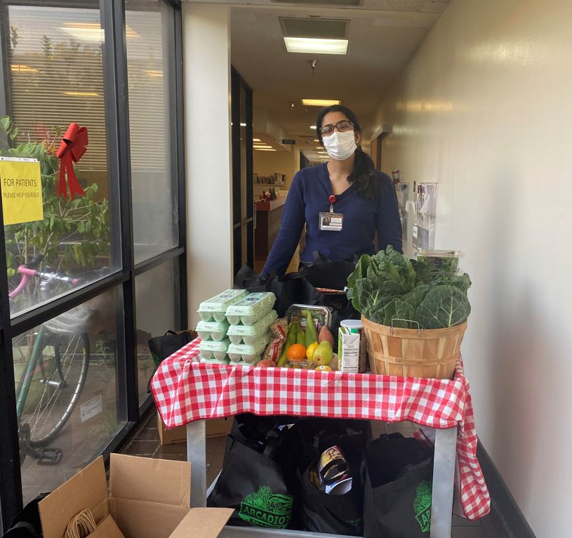 Saadhana standing behind a cart stacked with egg cartons, vegetables, and other food pharmacy items.